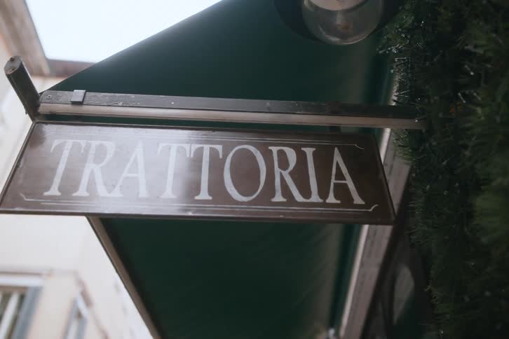 A brown sign with white lettering reading trattoria hangs beneath a green awning