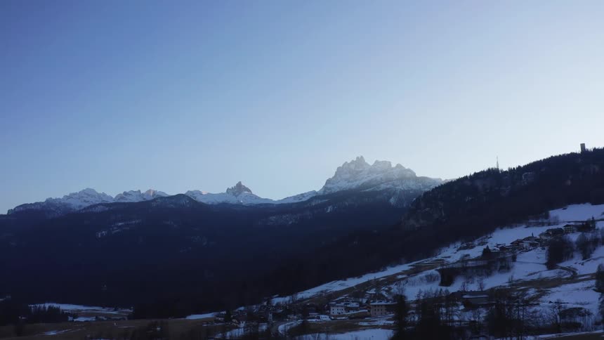 A distant mountain range with snowcapped peaks is silhouetted against a clear blue sky overlooking a dark forested valley and a few scattered buildings
