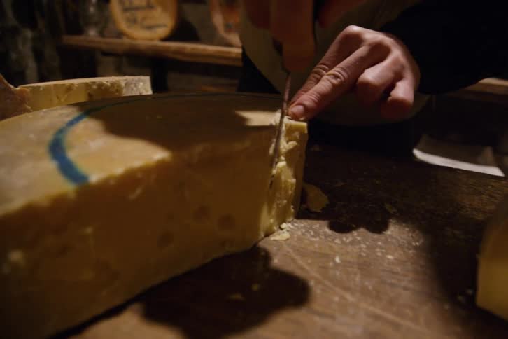 A persons hand uses a knife to cut a thick slice from a large block of cheese on a wooden surface