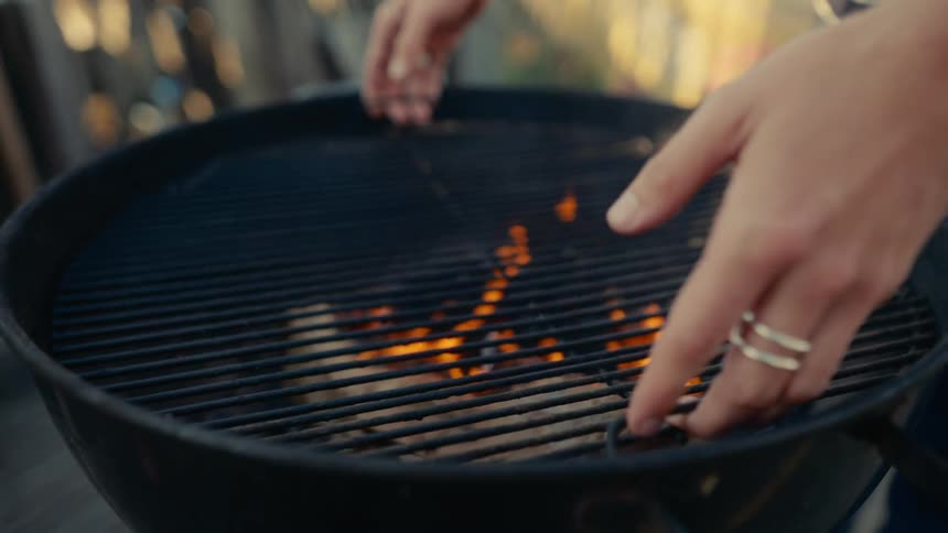 Aperson puts the cooking grate on a charcoal grill that has flaming charcoal