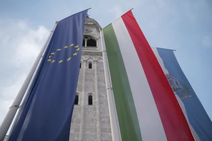 Three flags including the european union flag and the hungarian flag hang in front of a stone tower against a cloudy sky
