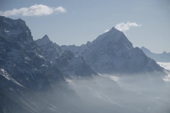 A series of jagged snowdusted mountain peaks rise through a layer of lowlying clouds under a bright sky