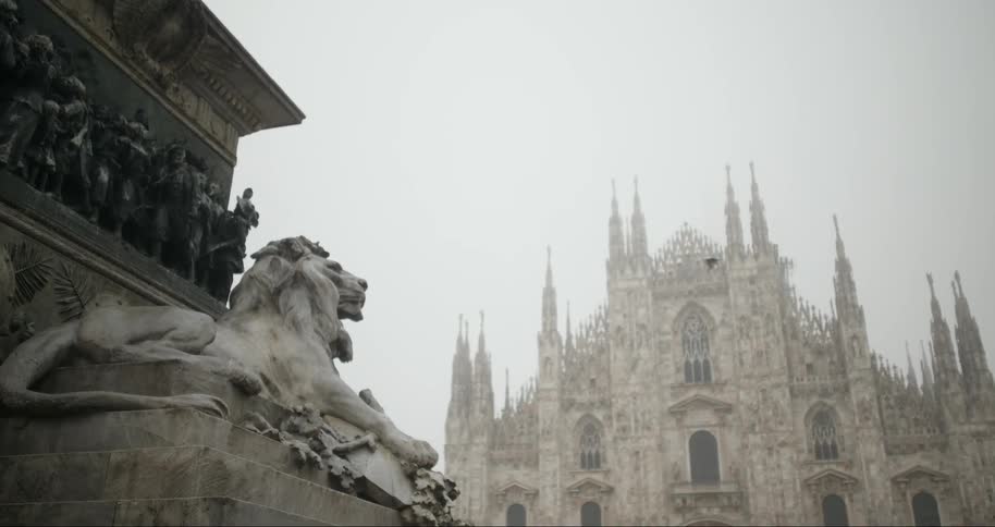 A marble lion statue stands prominently with the milan cathedral faintly visible in the foggy background