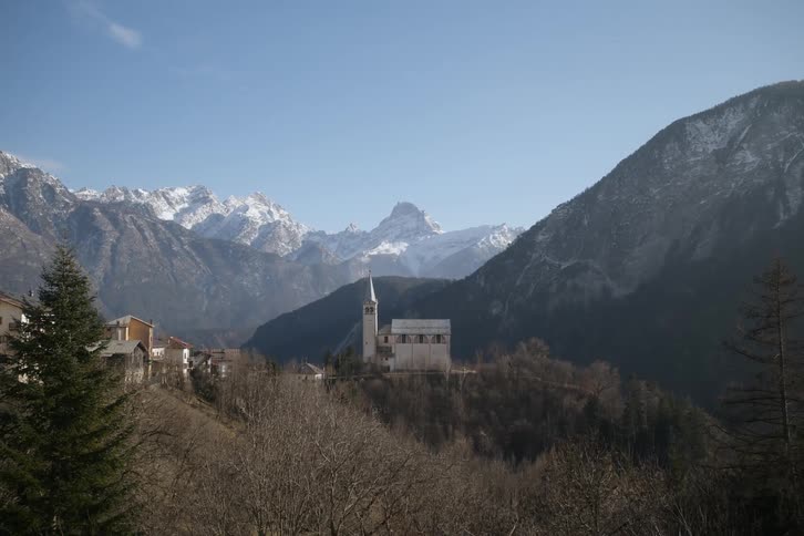 A small church with a steeple stands on a hilltop overlooking a village nestled in a valley surrounded by snowcapped mountains