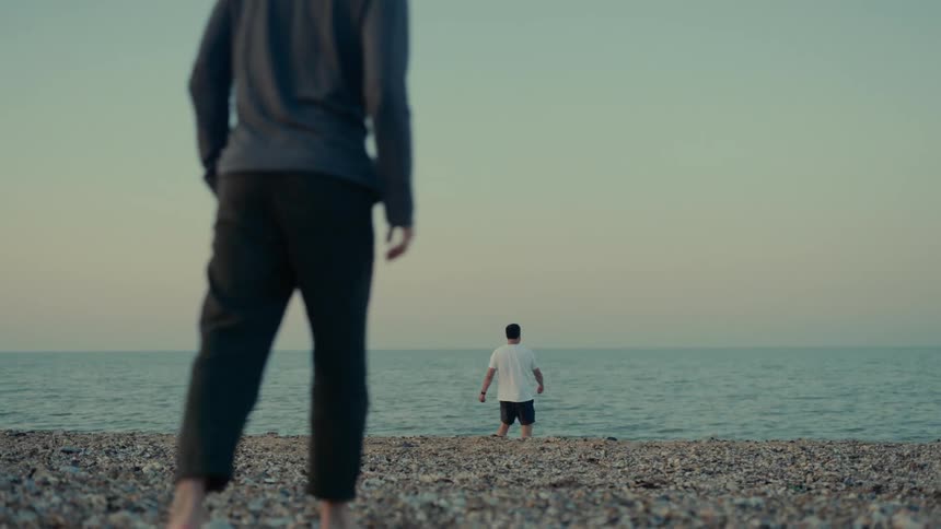 A person walks towards the shore of a pebbled beach where a man skips stones on the ocean
