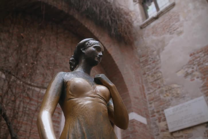 Bronze statue of shakespeares juliet set in a brickwalled courtyard with arches and dry vegetation in the background