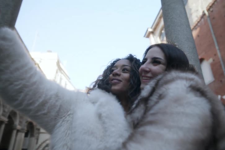 Two friends wearing fur coats are taking a selfie in front of a historic building with arched windows and a column