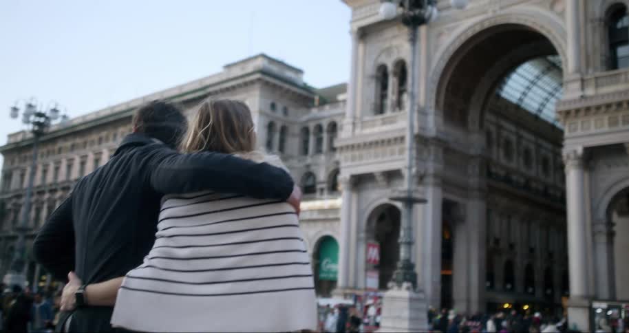 A man has his arm around a woman as they walk towards an arch on an old building