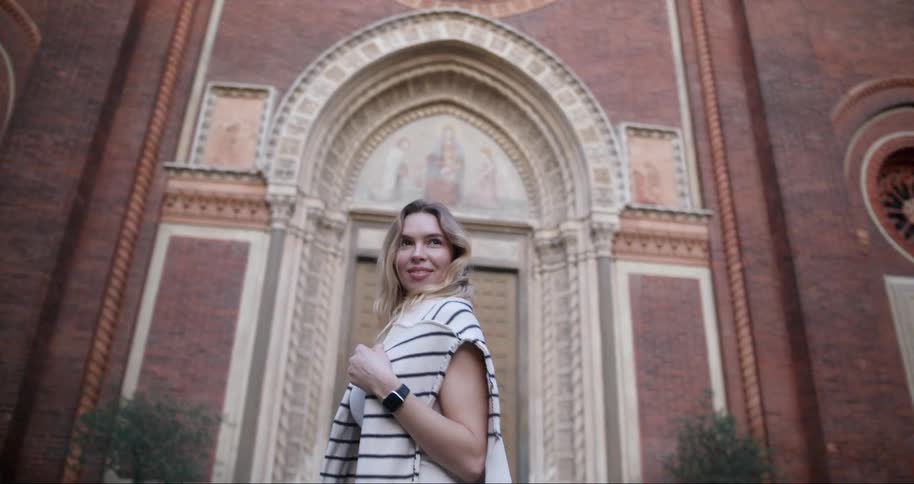 A woman with a striped sweater over her shoulders stands in front of the facade of an ornate brick building
