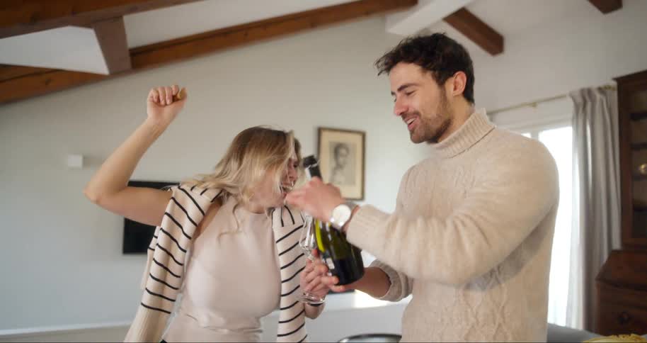 A man and woman smile as a woman pulls a cork from a bottle in a house