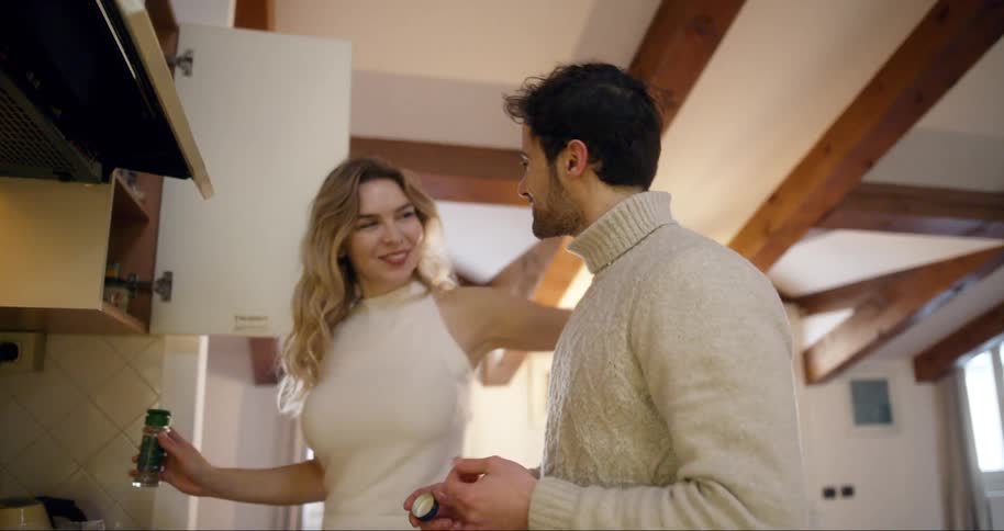 A woman holds a glass in a kitchen as a man blows a kiss
