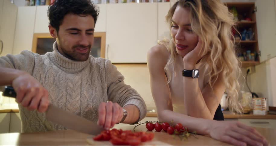A man slices tomatoes on a wooden cutting board in a modern kitchen while a blonde woman stands nearby watching
