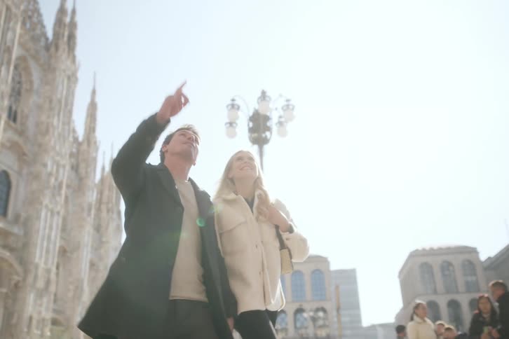 A smiling man and woman walk outdoors in bright sunlight with an ornate gothicstyle cathedral and a vintage street lamp in the background of a european city