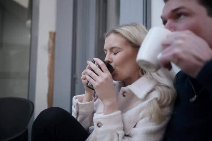 A young couple sits outdoors sitting closely together while drinking a hot beverage
