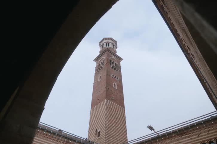 A tall brick clock tower of the parma cathedral with a decorative top is framed by two arched structures against a cloudy sky