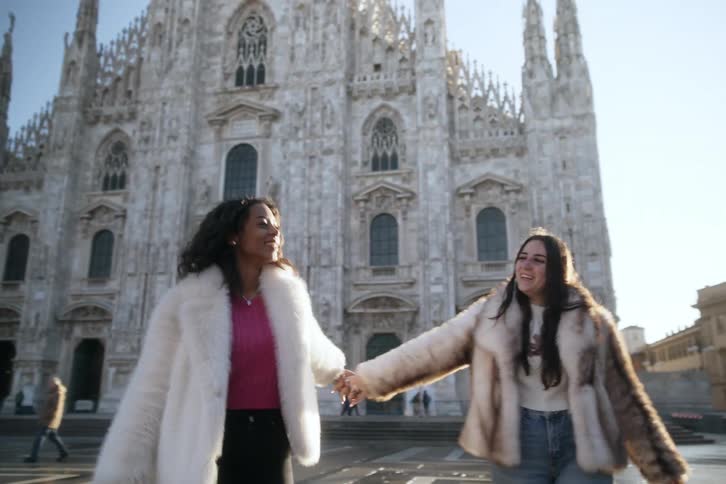 Two women smile and hold hands as they swing their arms and walk away from a gothic church