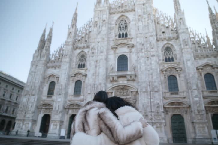 Couple wearing fur coats walks toward a cathedral arm in arm