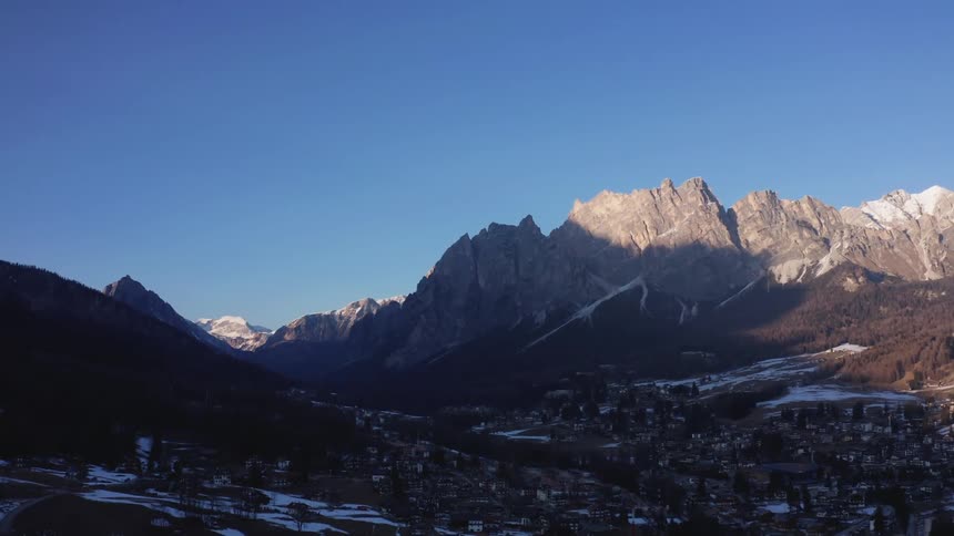 A wide shot of a mountain range with rocky peaks partially lit by sunlight casting shadows over a valley and a small town