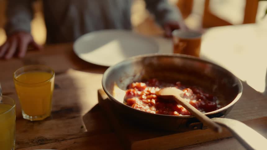 A person sits at a table with a skillet of tomatobased dish a glass of orange juice and an empty plate