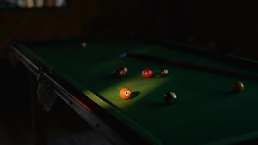 A pool table with several billiard balls scattered across its surface including an orange 5ball prominently highlighted by a beam of light