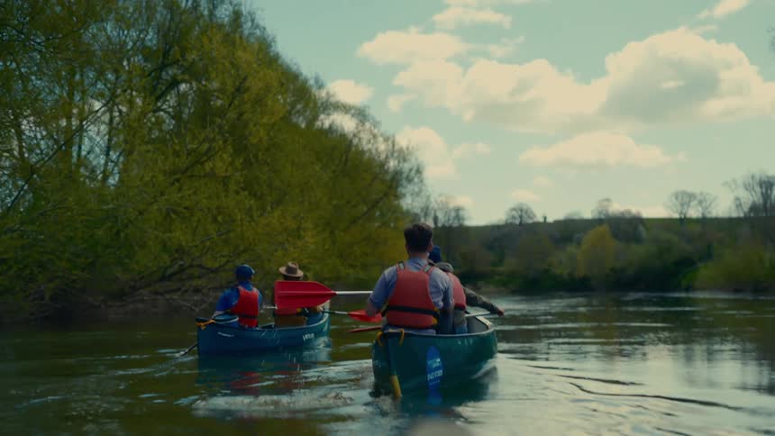 Two canoes with people wearing life vests paddle down a calm river on a bright partly cloudy day