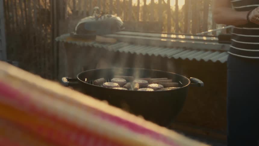Man walks away from an open grill at sunset while grilling burgers and franks