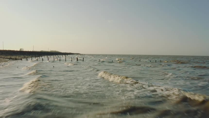 Waves wash around weathered wooden posts protruding from the shoreline stretching into the distance under a pale sky
