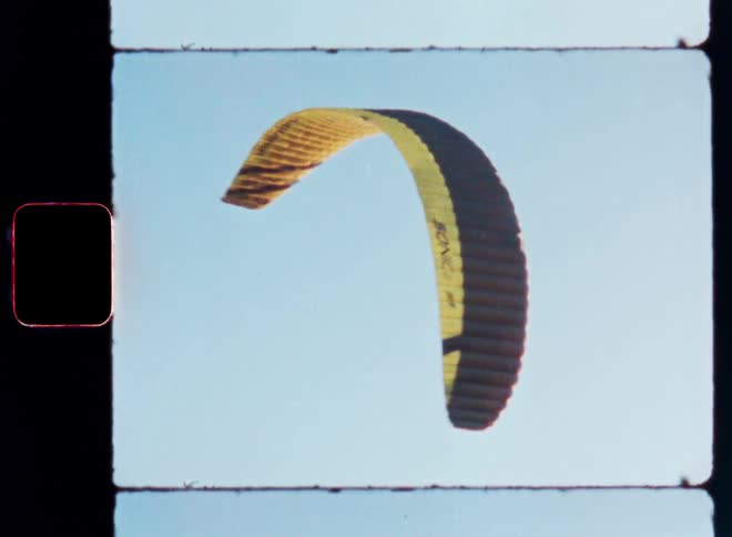 Within an analog film reel frame a fully deployed yellow and black wind chute dynamically rotates against a backdrop of clear blue sky capturing the essence of motion and form in vintage film style