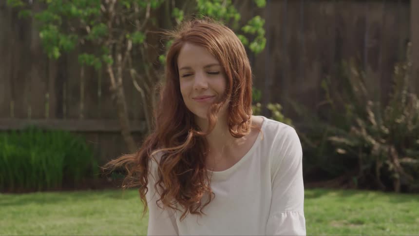 The face of a young redhaired woman with her hair blowing in the wind and she is wearing a subtle smile and a white outfit