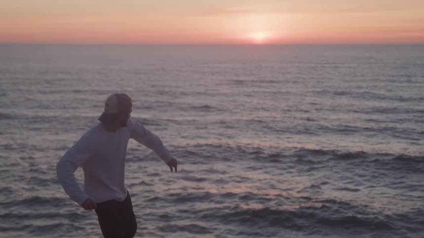 A man in a cap and white shirt is standing facing the sea and then he throws something very long