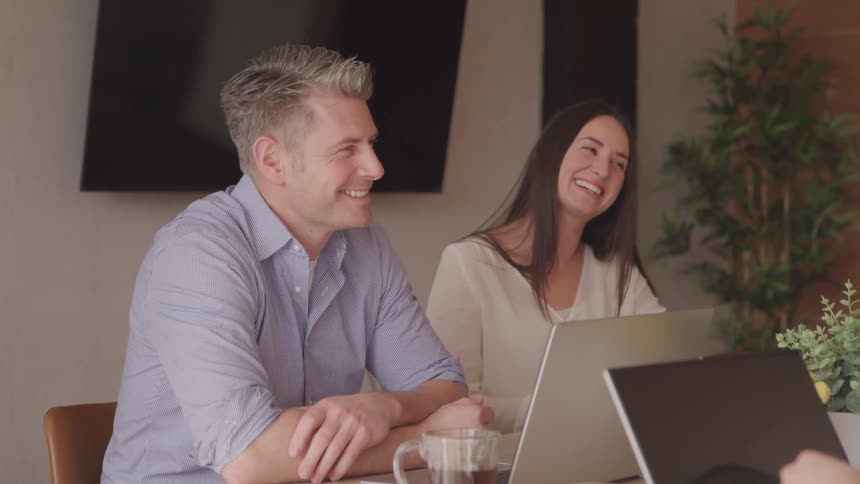 A smiling man and woman sit at a table with laptops and a cup of tea both looking off to the side