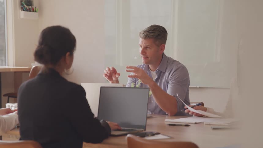 A man gestures while speaking at a table with other people one using a laptop during a meeting