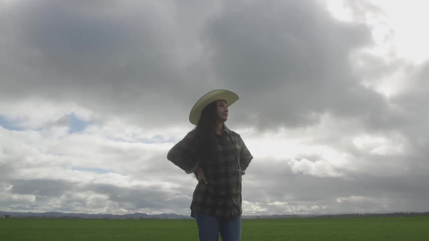 A woman in a cowboy hat and plaid shirt stands in a green field with her hands on her hips looking towards the horizon under a cloudy sky