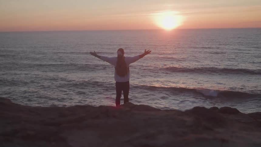 Traveler spreading his arms while standing on a rock by the sea with the sunrise on the horizon