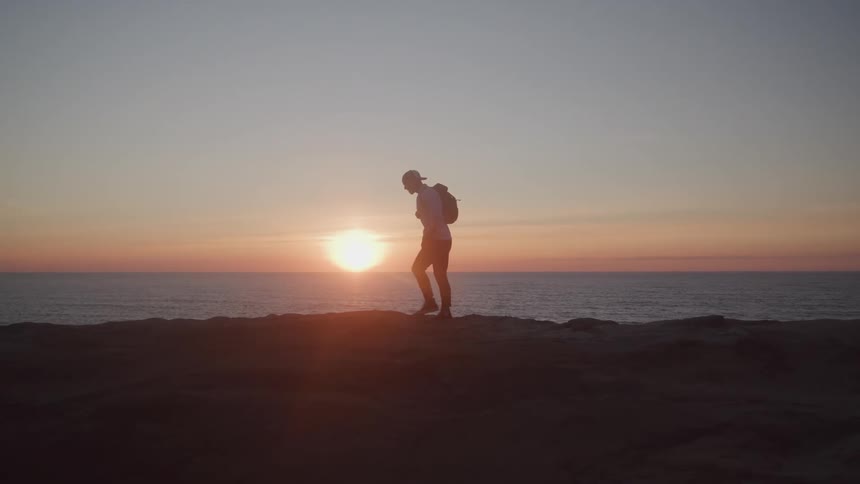 Silhouette of a traveler with a backpack walking on a rock by the sea with the sunrise on the horizon