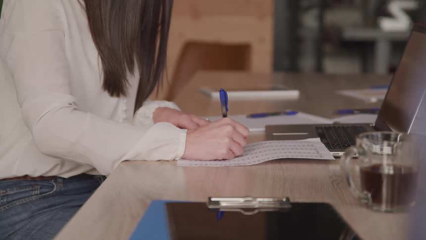 The hand of a person who is wearing white clothes and is writing with a pen on a paper that is on top of a wooden table