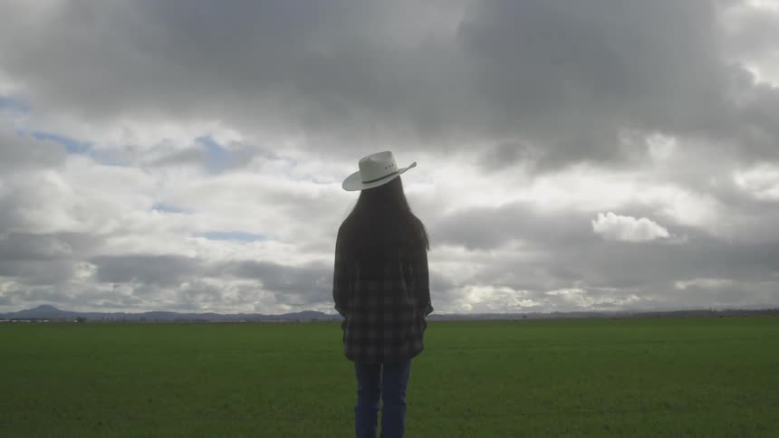 Woman with her back in a green field while wearing a hat on a cloudy day