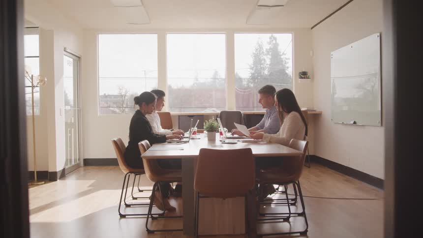 A diverse group of business professionals are seated around a table in a brightly lit office working on laptops and papers during a meeting