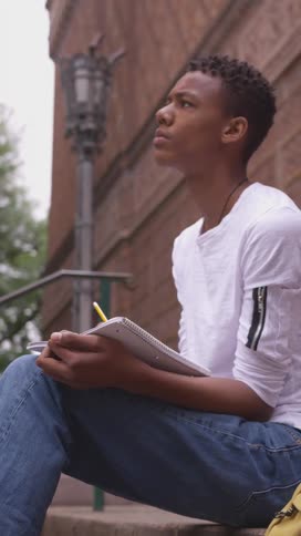 Student sitting on the outside stairs of a building writing in a notebook and becoming thoughtful