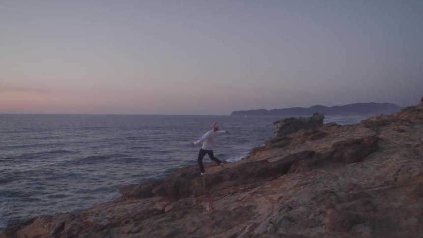 A man stands on a rocky cliff and throws a small object into the ocean at sunset