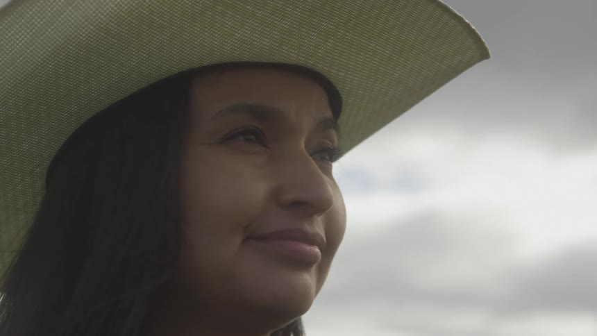 A woman in a cowboy hat gazes upward with a subtle smile against a cloudy sky