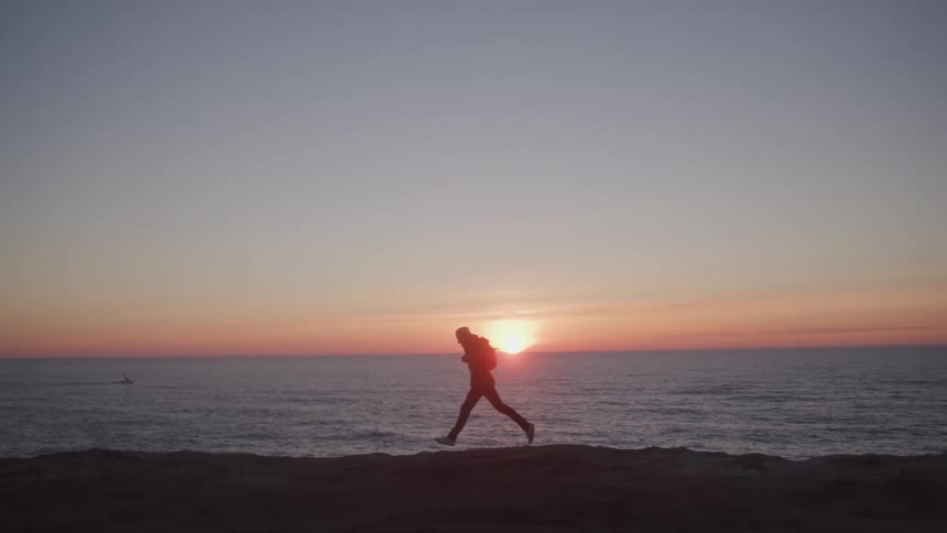 A silhouetted figure with a backpack runs along a coastline at sunset with a boat visible in the ocean