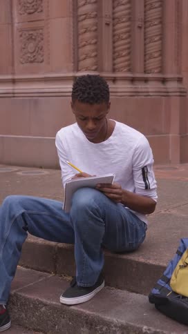Student sitting on the outside stairs of a building writing in a notebook
