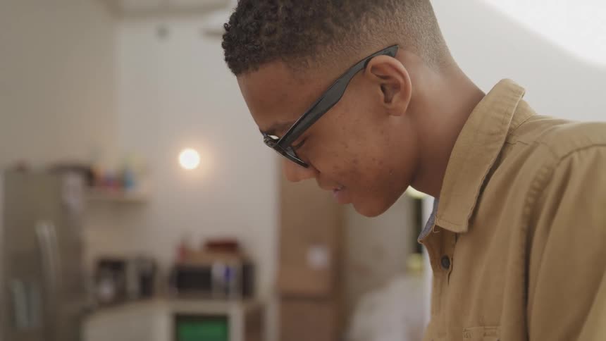 A young black man with short hair and glasses wearing a tan shirt looks down intently in an indoor space