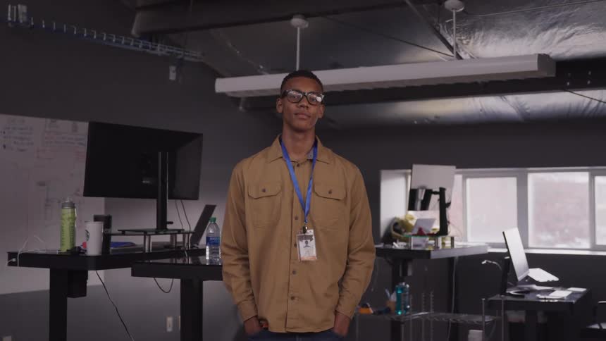 A young bespectacled black man with short hair wearing a tan shirt and employee id stands facing forward in a modern minimalist office space