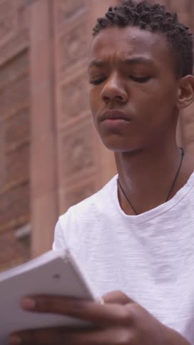 A young focused black person with short hair wearing a white shirt and necklace alternates between looking at a notebook and upwards in front of a brick building