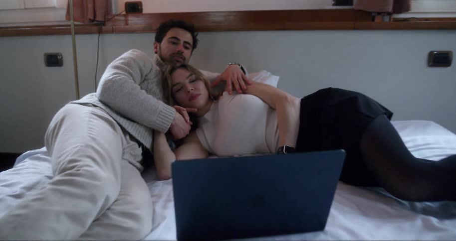 A young man and a woman are lying on a bed and the man is stroking the womans arm and they are both watching something on a laptop