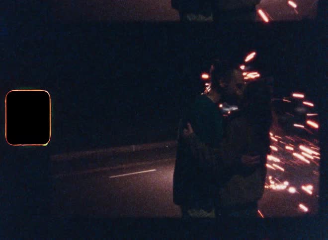Man and woman kiss while standing outdoors at night near sparking fireworks