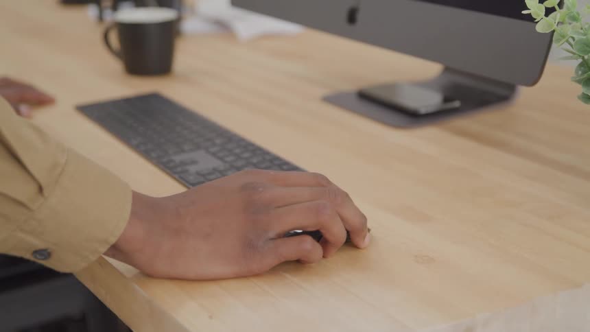 The hand of a person wearing a longsleeved garment moving a mouse that is on a wooden table in front of a computer screen