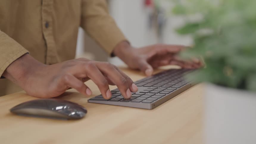A person types on a black keyboard placed on a wooden desk with a green plant and a computer mouse visible nearby